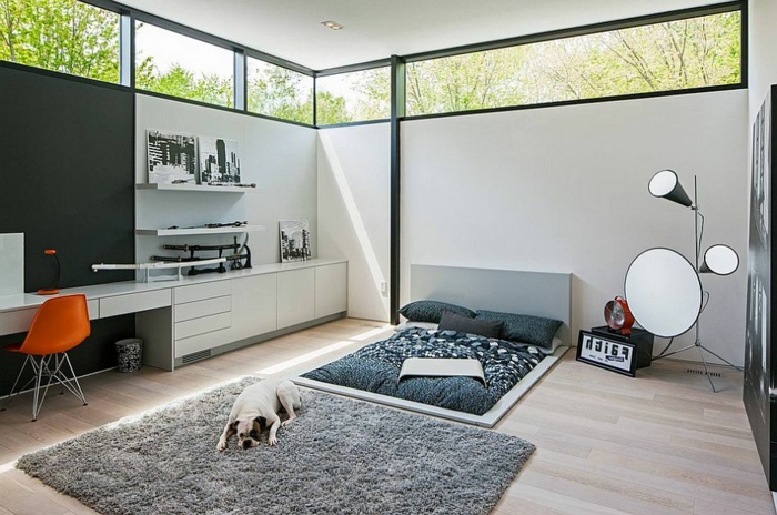 moderner Einrichtun in skandinavischem Stil - niedriges Bett mit grauer Bettwäsche, Stehlampe mit zwei Birnen und einem runden Spiegel, Fenster hoch an der Decke, Led-Beleuchtung, oranger Stuhl aus Plastik, ein Hund liegt auf dem Boden